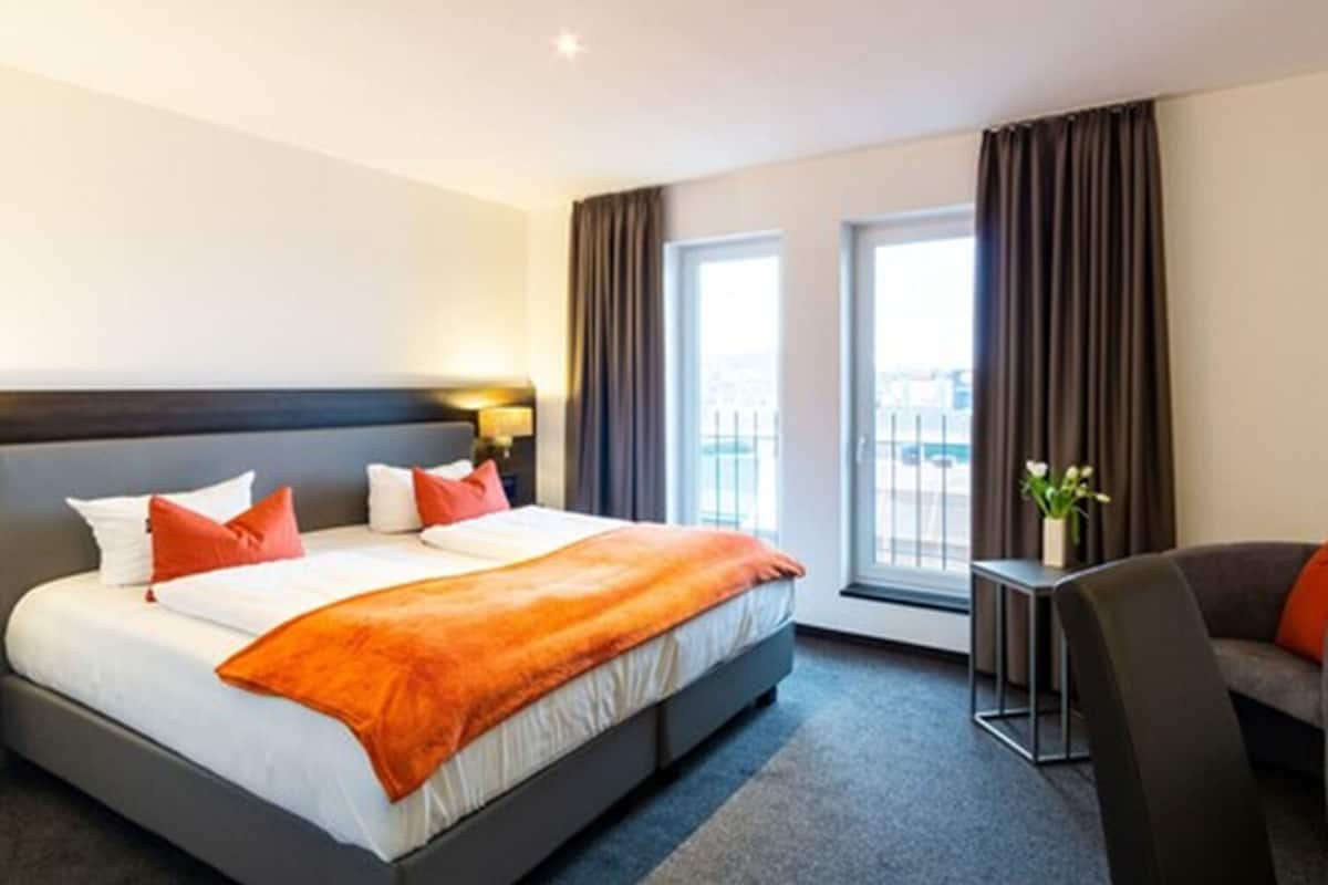 A hotel room with a large bed, a desk, and a chair. There are two windows with curtains.