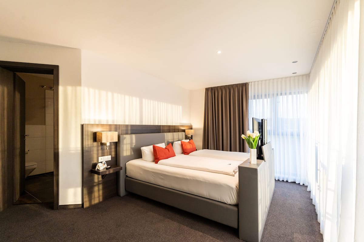 A modern hotel room with a large bed, bedside tables, a mirror, and a view of the outdoors through sheer curtains.