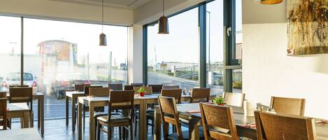 A modern dining area with wooden tables and chairs, large windows, and pendant lighting.
