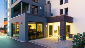 Un hotel moderno con fachada de vidrio, zona de estar al aire libre y un restaurante visible en su interior.