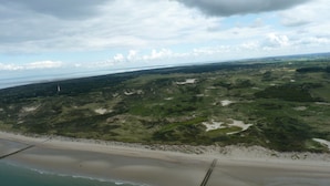 Vista aérea de un paisaje costero con una playa arenosa, vegetación verde y un faro a lo lejos.