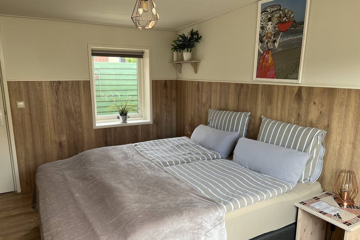 Een slaapkamer met een groot bed, houten lambrisering, een raam met een plant, een wandplank en een decoratief schilderij.