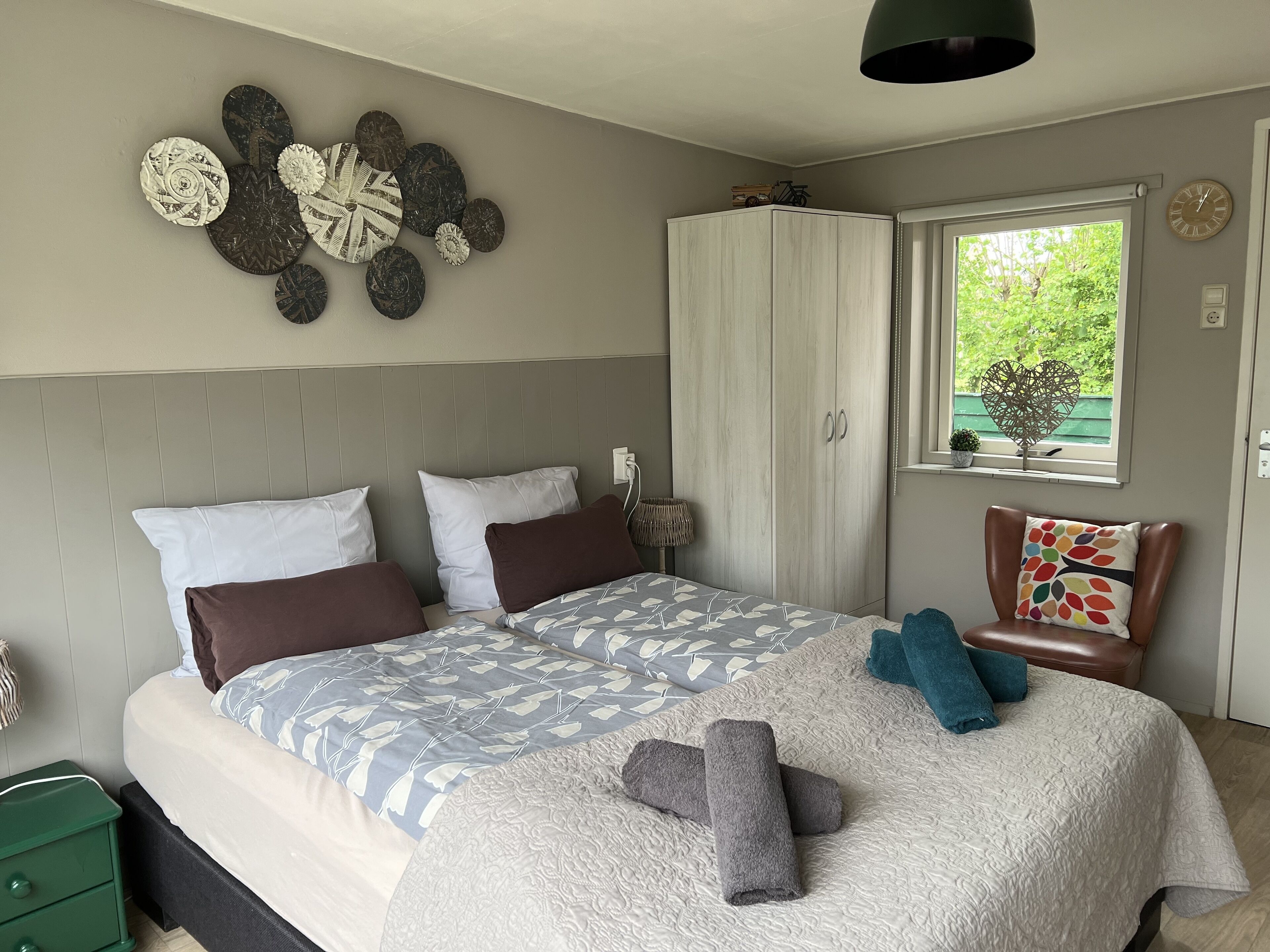 A bedroom with a bed, pillows, a wardrobe, a window with a view, and a wall decoration.