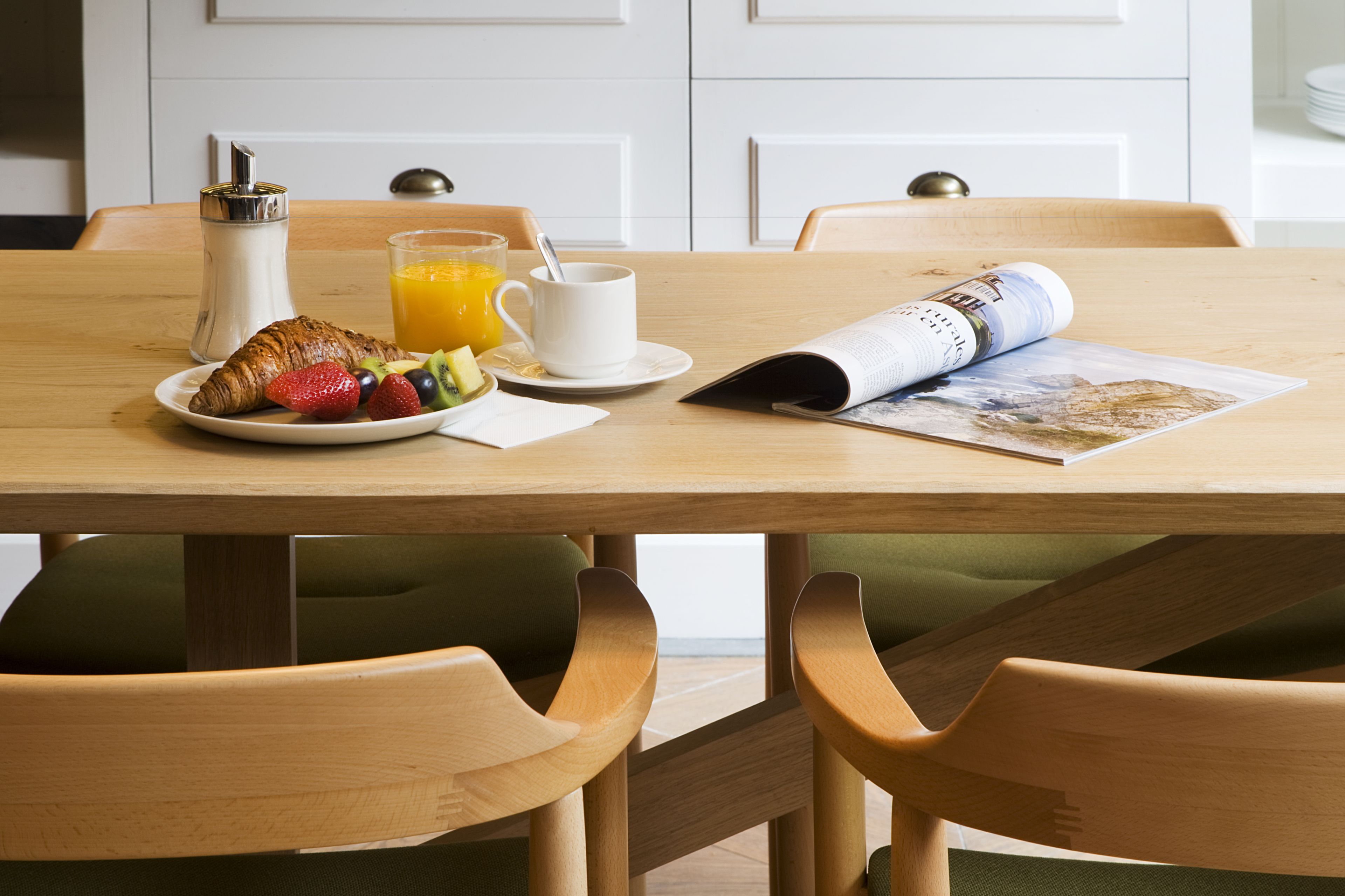 Une table de petit-déjeuner avec des croissants, des fruits, du jus d’orange et un journal.