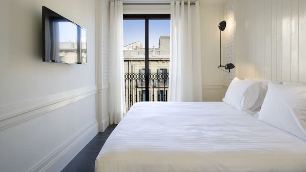 Habitación de hotel con cama, televisión montada en la pared, balcón con barandilla y ventana con cortinas.