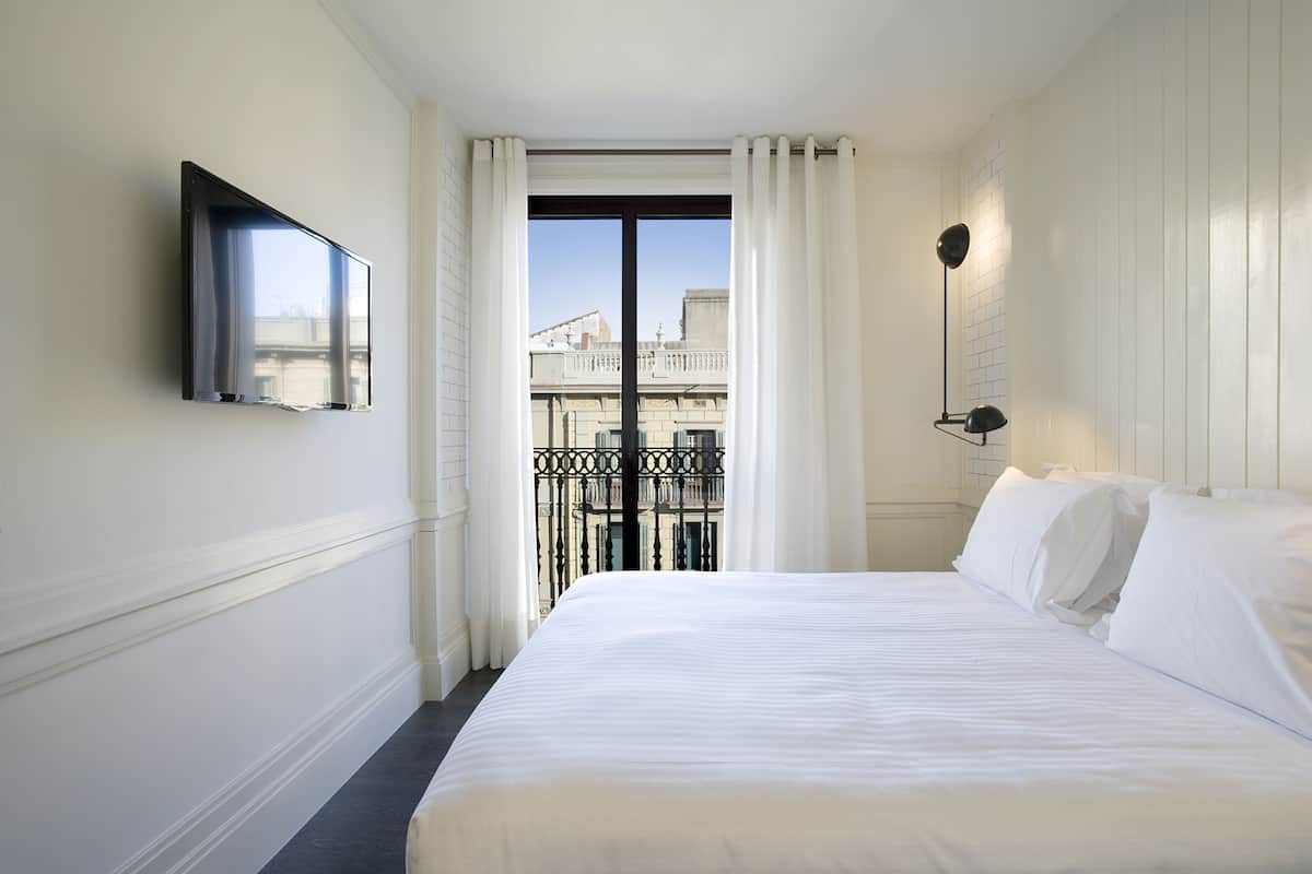 A hotel room with a bed, a TV mounted on the wall, a balcony with a railing, and a window with curtains.