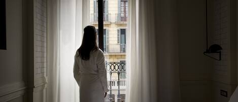 Una habitación con cama, ventana con cortinas y vista a un edificio con balcones.