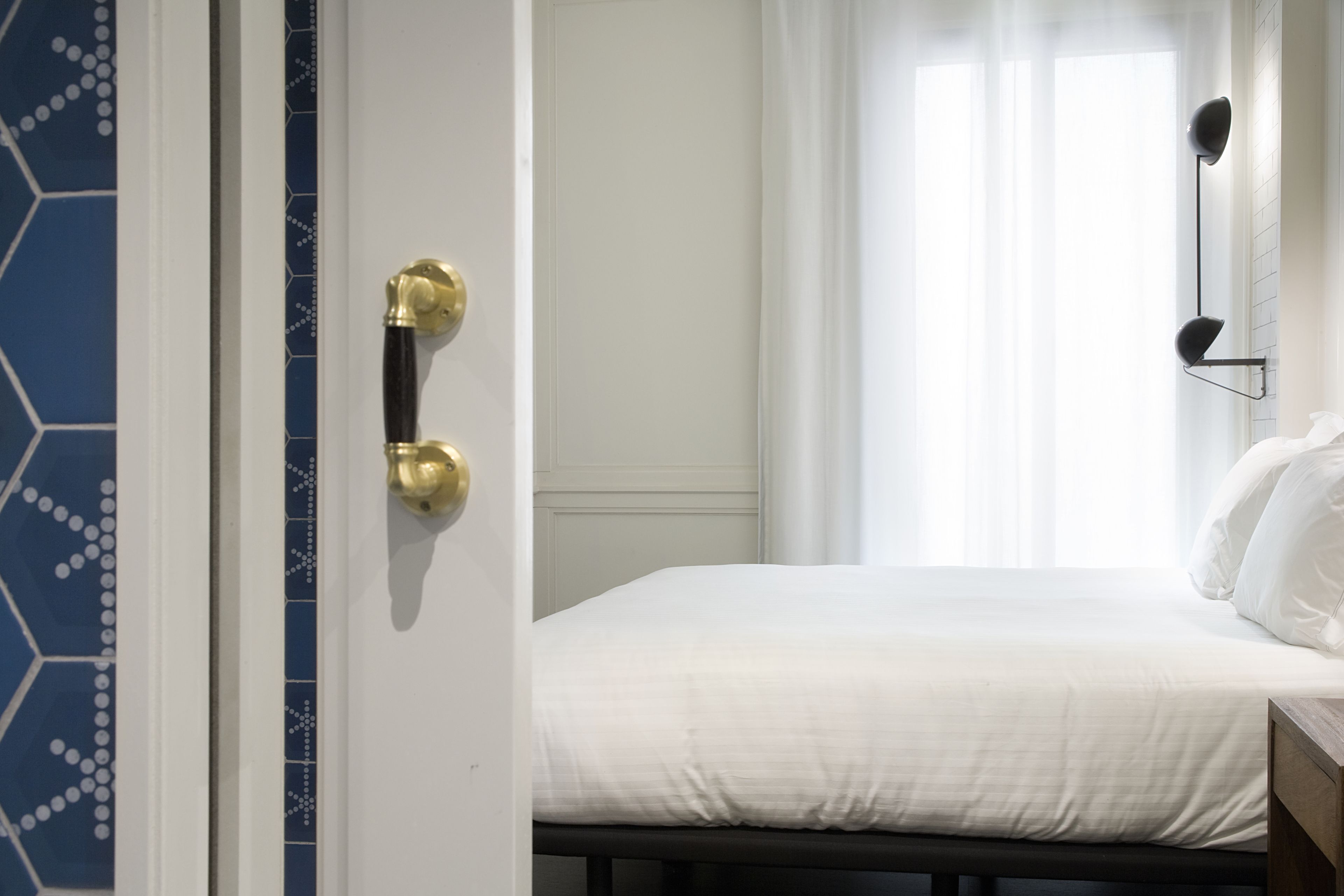 A hotel room with a bed, a bedside table, a wall-mounted lamp, and a door with a brass handle.