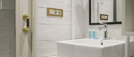 Un baño con lavamanos blanco, un espejo y una ducha. Hay dos enchufes en la pared.