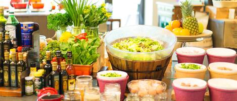 A variety of food items and condiments on a table, including jars of olives, bottles of olive oil, and a wooden bowl with greens.