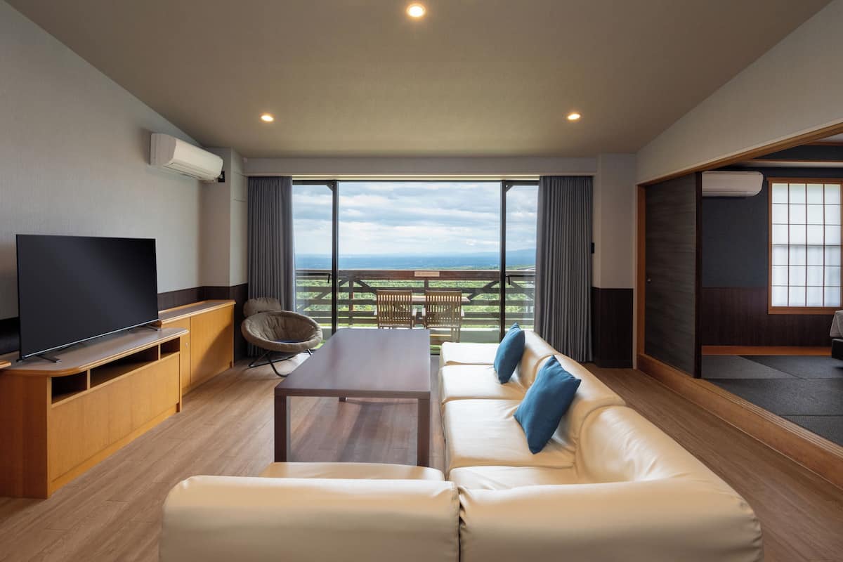 A modern living room with a flat-screen TV, wooden furniture, a sofa with blue cushions, and a balcony with a view of the countryside.