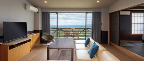 A modern living room with a flat-screen TV, wooden furniture, a sofa with blue cushions, and a balcony with a view of the countryside.