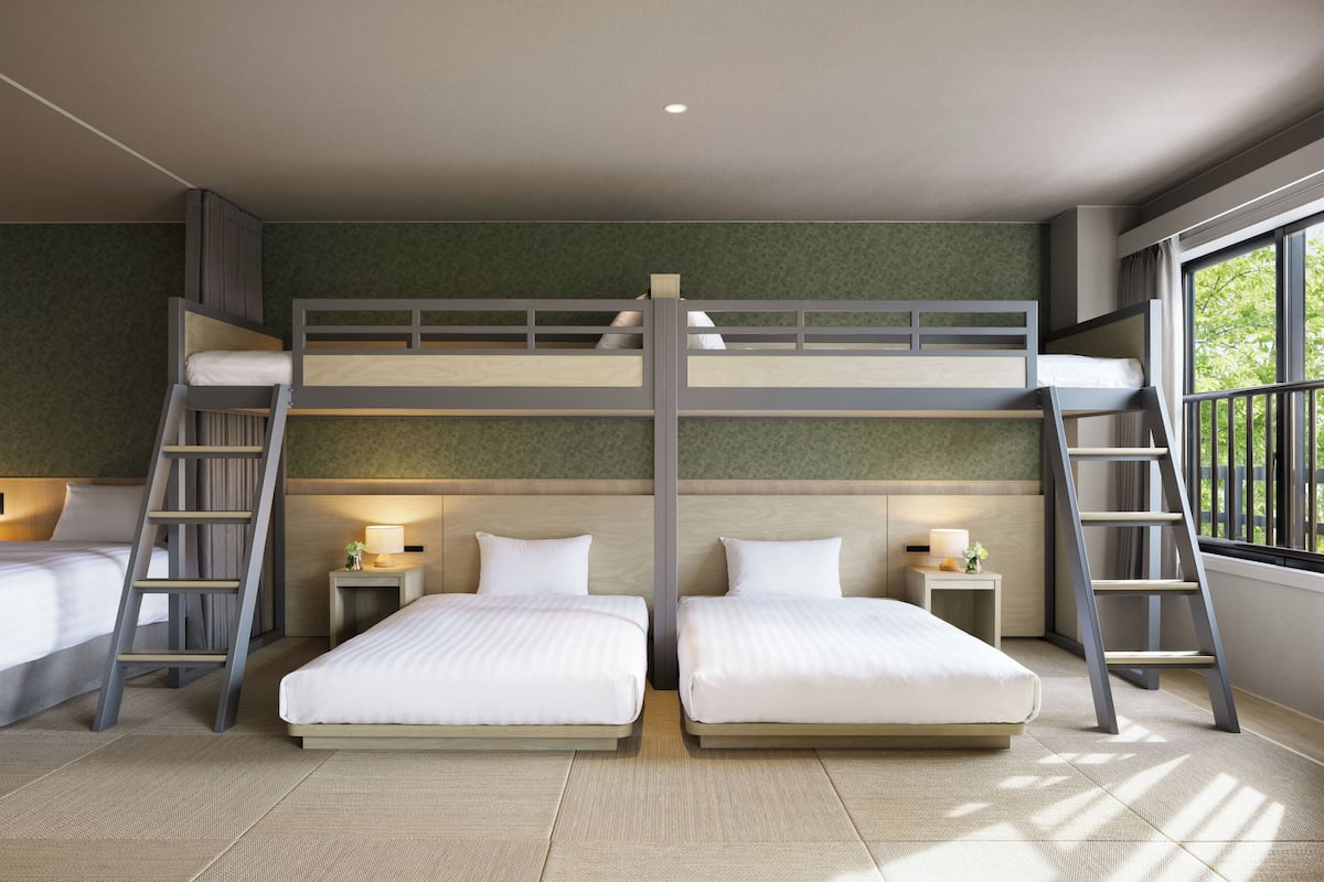 A room with bunk beds, two bedside tables with lamps, and a window with a view of trees.