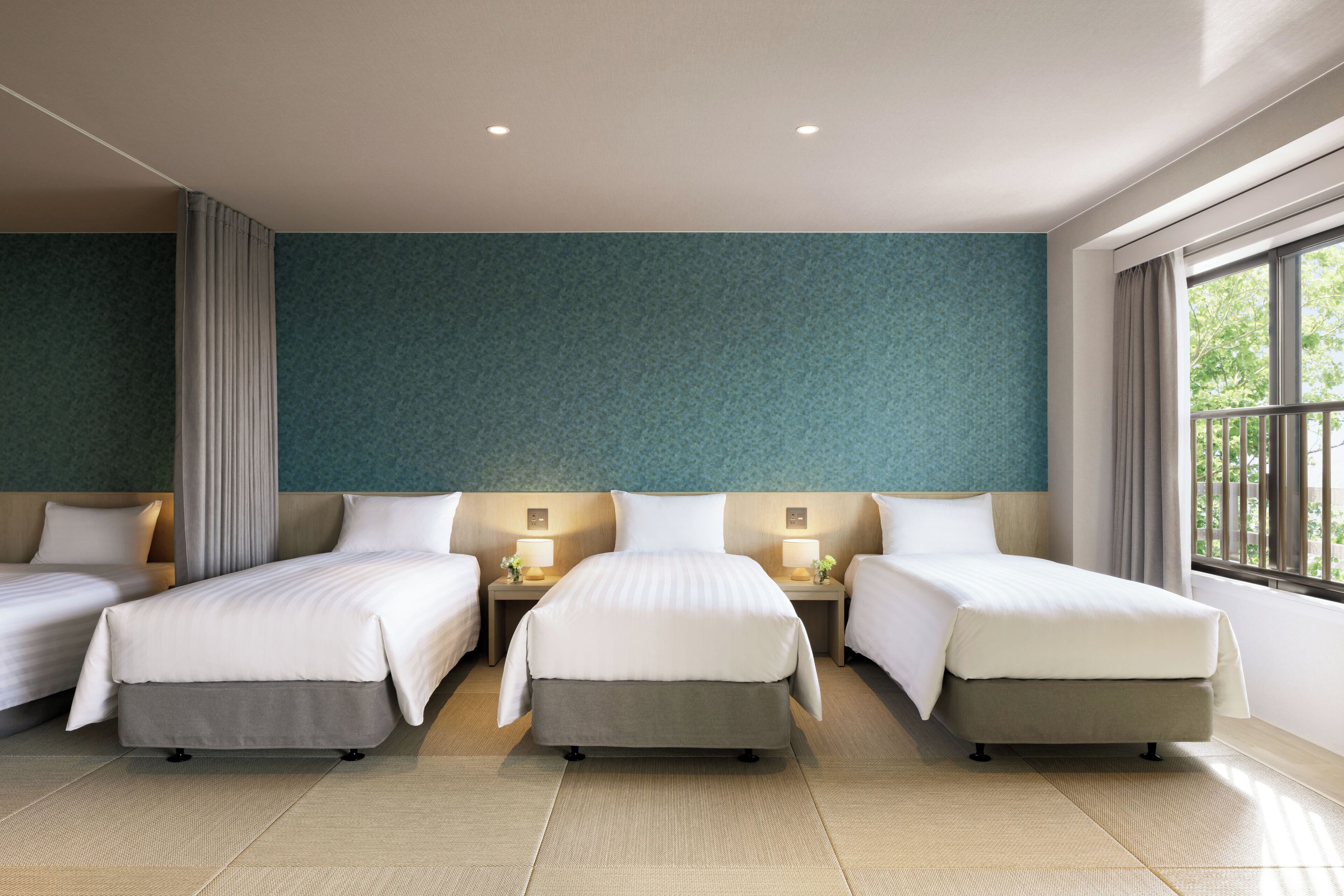 A hotel room with three beds, a large window, and a teal-colored wall.