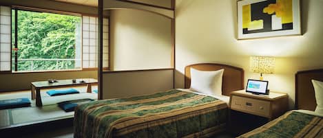 A hotel room with two beds, a desk, a chair, a small table, a nightstand with a lamp, and a window with a view of greenery.