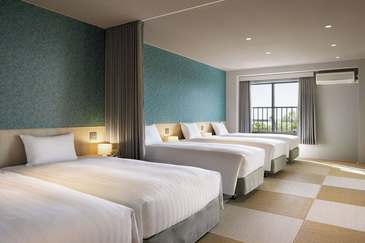 A hotel room with three beds, a large window with curtains, and a view of greenery outside.