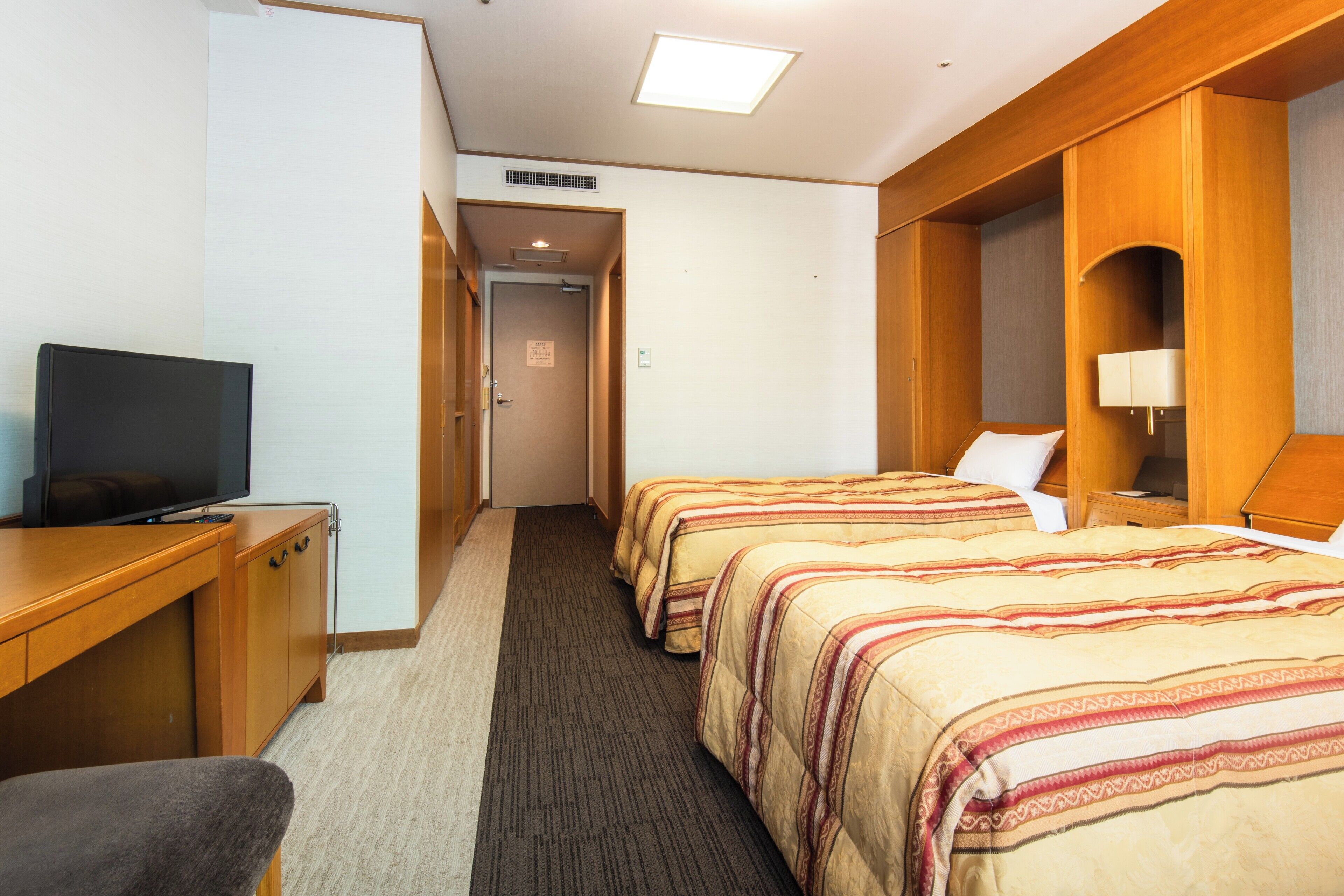 Une chambre d’hôtel avec un téléviseur posé sur un bureau, deux lits avec une literie à rayures, un sol recouvert de moquette et des panneaux de bois au mur.