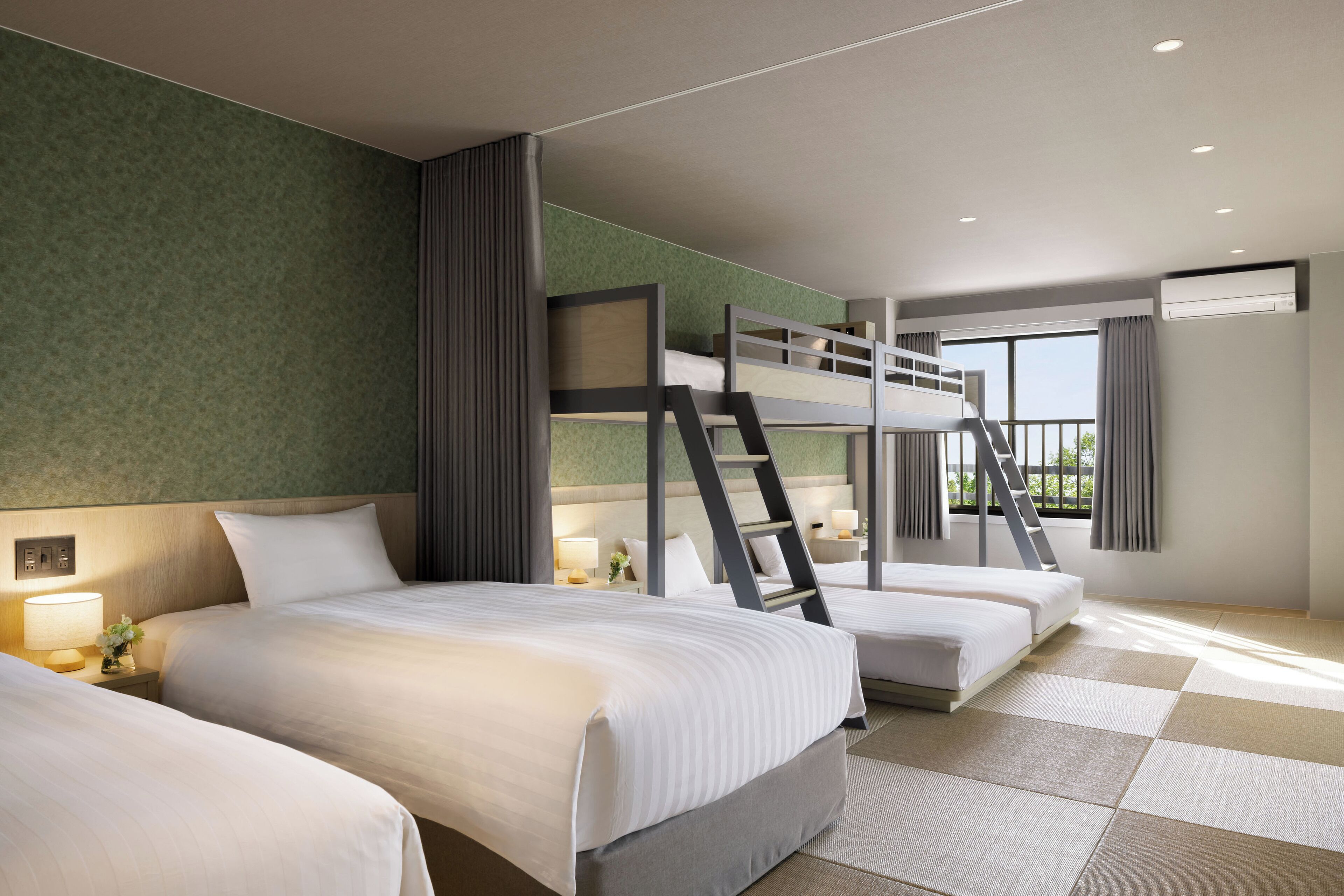 Une chambre d’hôtel avec deux lits, un lit superposé, une fenêtre avec des rideaux et une petite table avec une lampe.