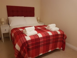 Een netjes opgemaakt bed met een rood-wit geruit plaid, witte kussens en twee witte nachtkastjes met lampen.