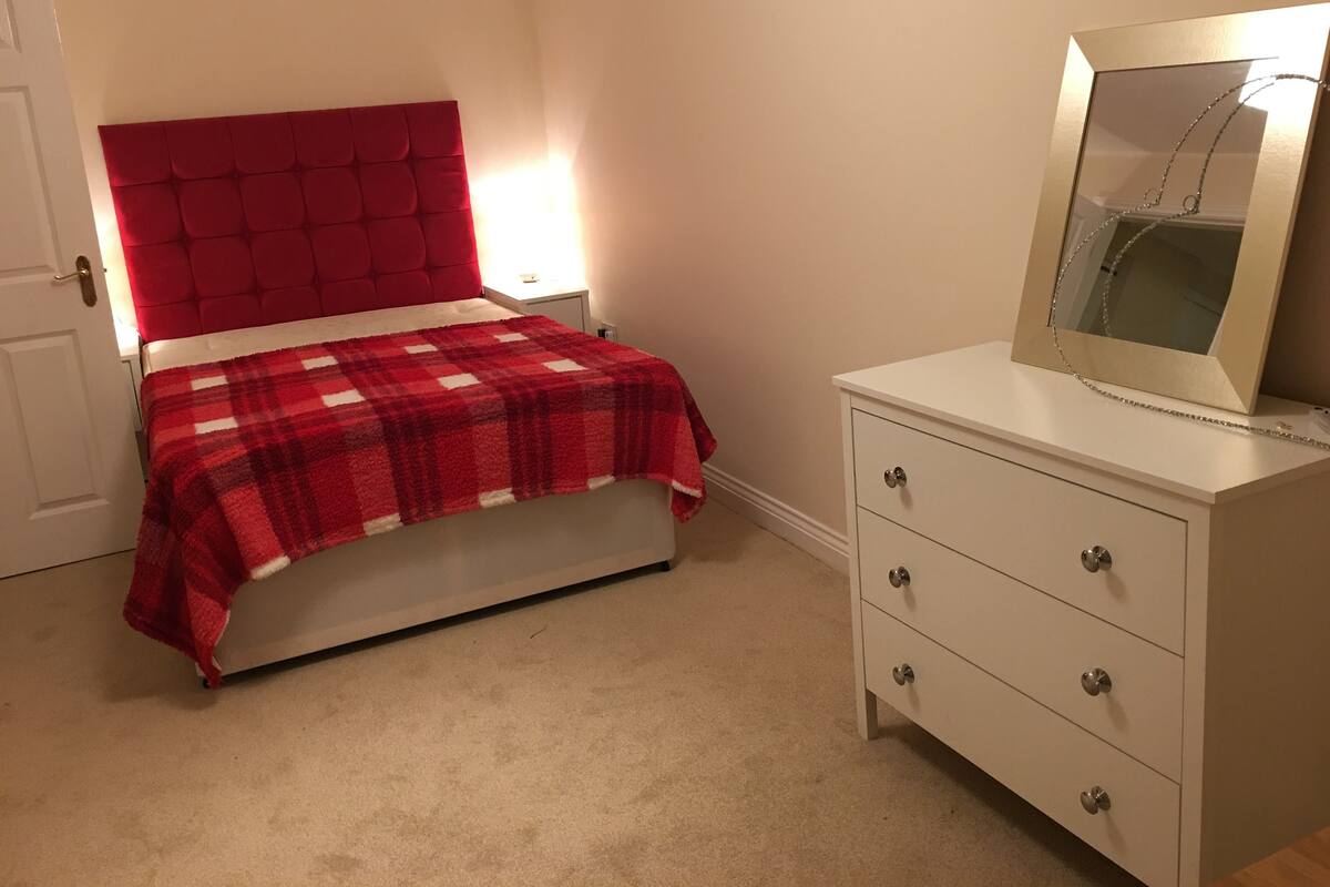 A bedroom with a red tufted headboard, a plaid bedspread, a white dresser with drawers, and a mirror with a chain.