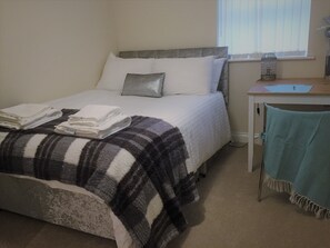 A neatly made bed with white linens and a plaid blanket, a desk with a laptop and a vase, and a window with blinds. - Genesis Home Serviced Apartment (Coventry)