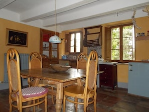 Uma sala de jantar com mesa e cadeiras de madeira, um lustre e vista para a cozinha através de uma porta aberta.