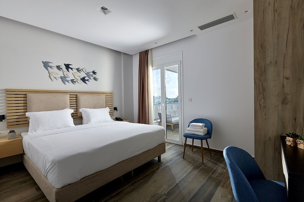 Moderni hotellihuone, jossa on suuri sänky, puinen pääty, yöpöytä, sininen tuoli ja ikkunasta avautuva näkymä kaupunkiin.