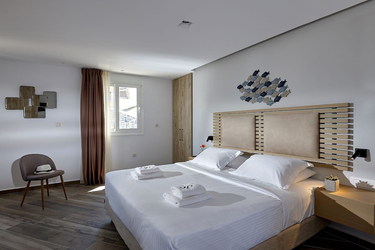 Moderni hotellihuone, jossa on suuri sänky, puinen pääty, seinään kiinnitetty valaisin ja ikkunasta avautuva näkymä ulos.