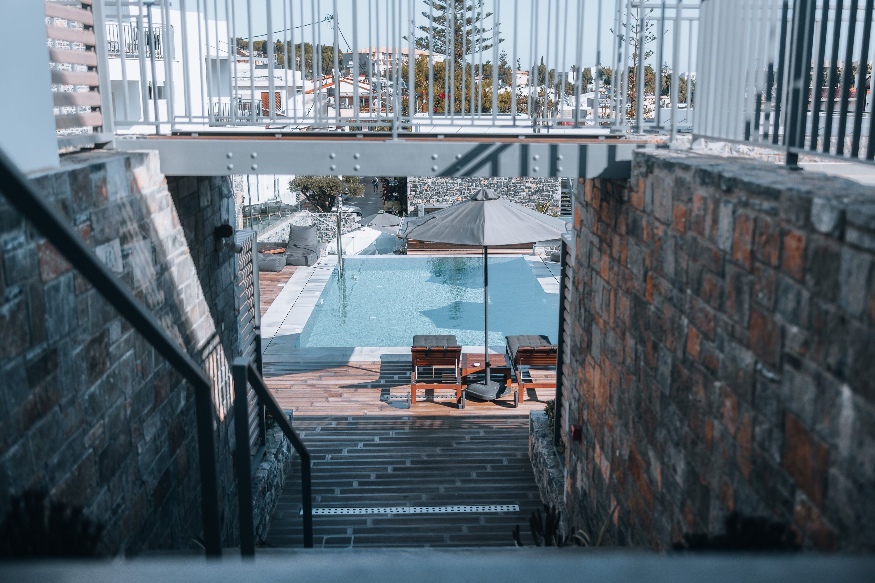 Udsigt til en swimmingpool med liggestole og et pergolaområde, set fra en trappe med murstensvægge.