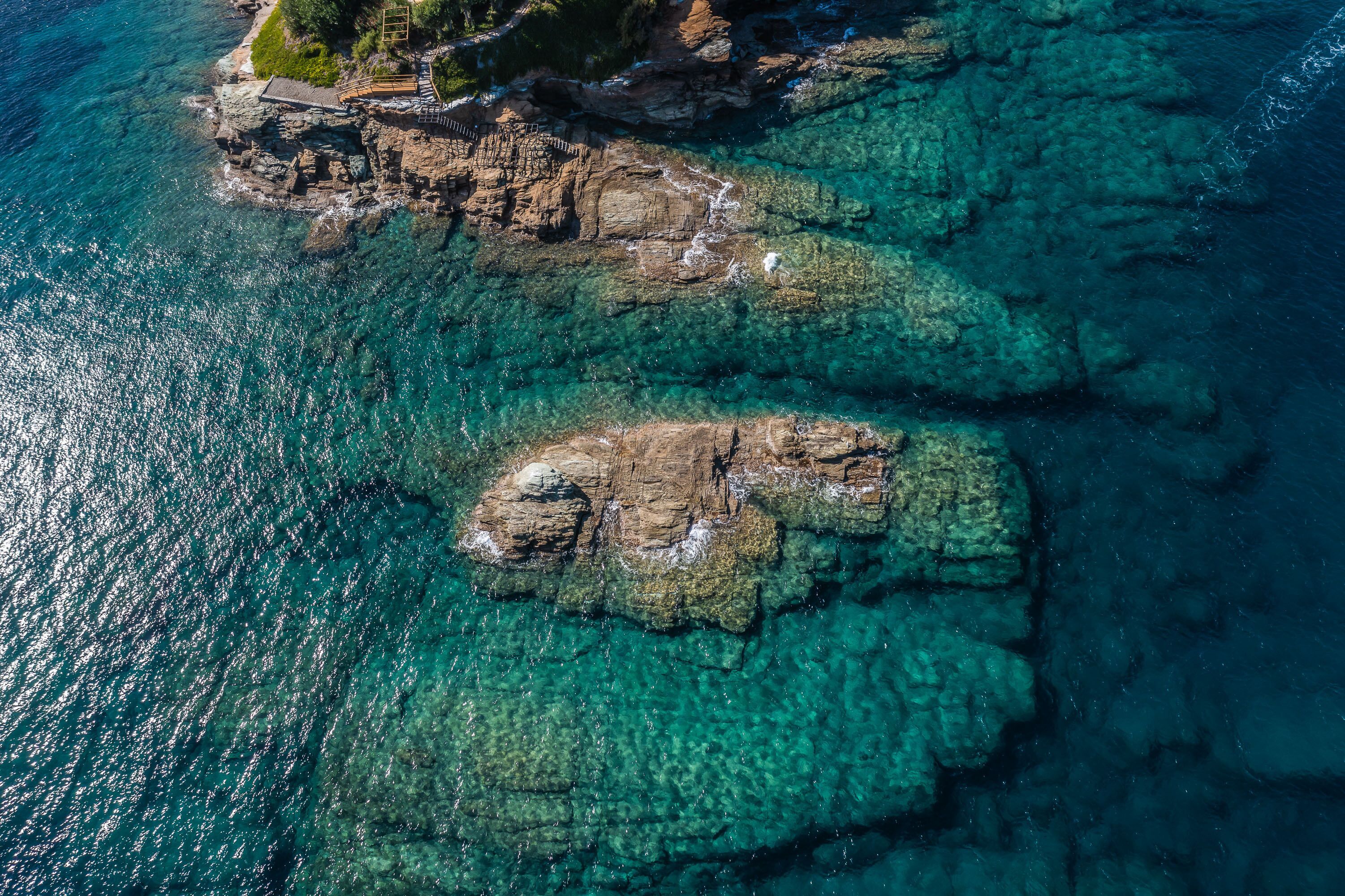 An aerial view of a coastal area with clear turquoise waters, rocky formations, and a small pathway leading to a grassy area.