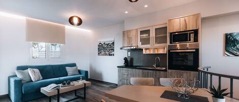 A modern living room with a sofa, coffee table, and dining table. There is a kitchen area with cabinets and appliances.