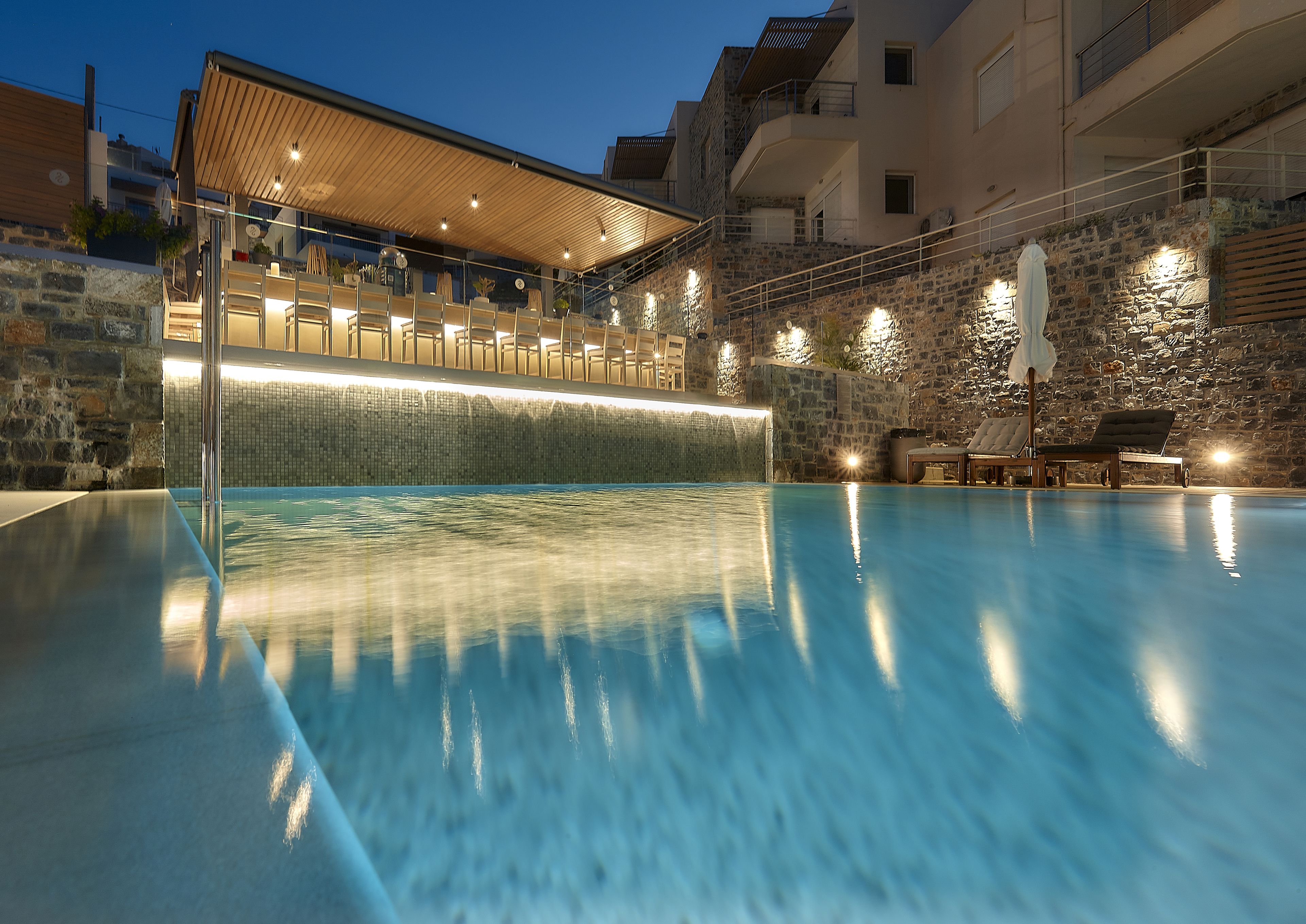 Une piscine dotée d’une terrasse en bois et de murs en pierre, entourée d’espaces de détente en plein air et d’une zone couverte éclairée.