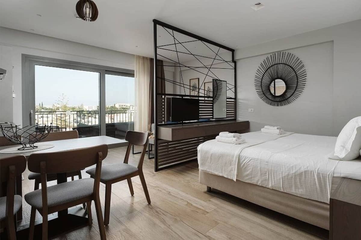 Habitación de hotel moderna con una cama, un escritorio con sillas, un ventanal grande, un televisor y un elemento decorativo en la pared.