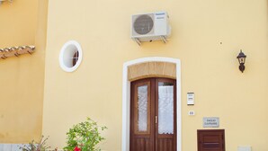 Un edificio con fachada amarilla, puerta de madera con paneles de vidrio, unidad de aire acondicionado pequeña y plantas en macetas.