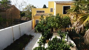 Un edificio amarillo con techo de tejas, rodeado de vegetación y un muro blanco.