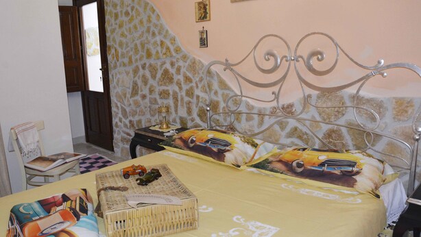 Un dormitorio con pared de piedra, cama con cabecero metálico, mesita de noche con lámpara y una silla con cojín.