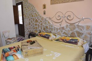 Quarto com parede de pedra, cama com cabeceira de metal, mesa de cabeceira com luminária e uma cadeira com almofada.