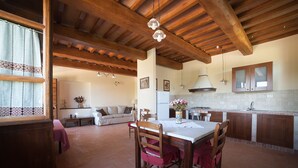 A spacious living area with a wooden ceiling, a dining table with chairs, a kitchen with wooden cabinets, and a sofa in the background. - Agriturismo Il Beccafico Alpaca (Gubbio)