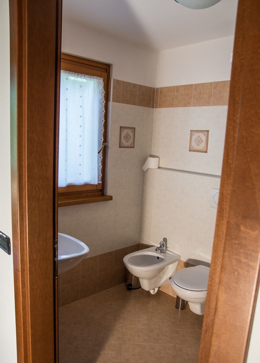 Une petite salle de bain avec un lavabo, un bidet et des toilettes. Il y a une fenêtre avec des rideaux en dentelle et un cadre photo accroché au mur.