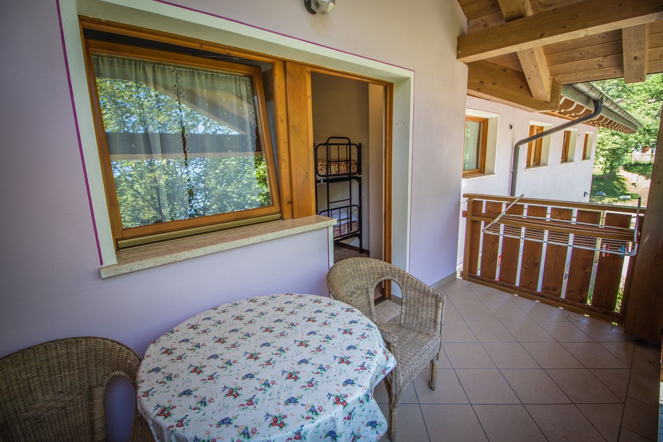 A room with a round table covered in a floral tablecloth, two wicker chairs, a window with a curtain, and a view of a balcony with a railing.