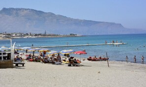 Une plage avec des chaises longues et des parasols, une jetée qui s’avance dans l’eau, et des gens qui se baignent.