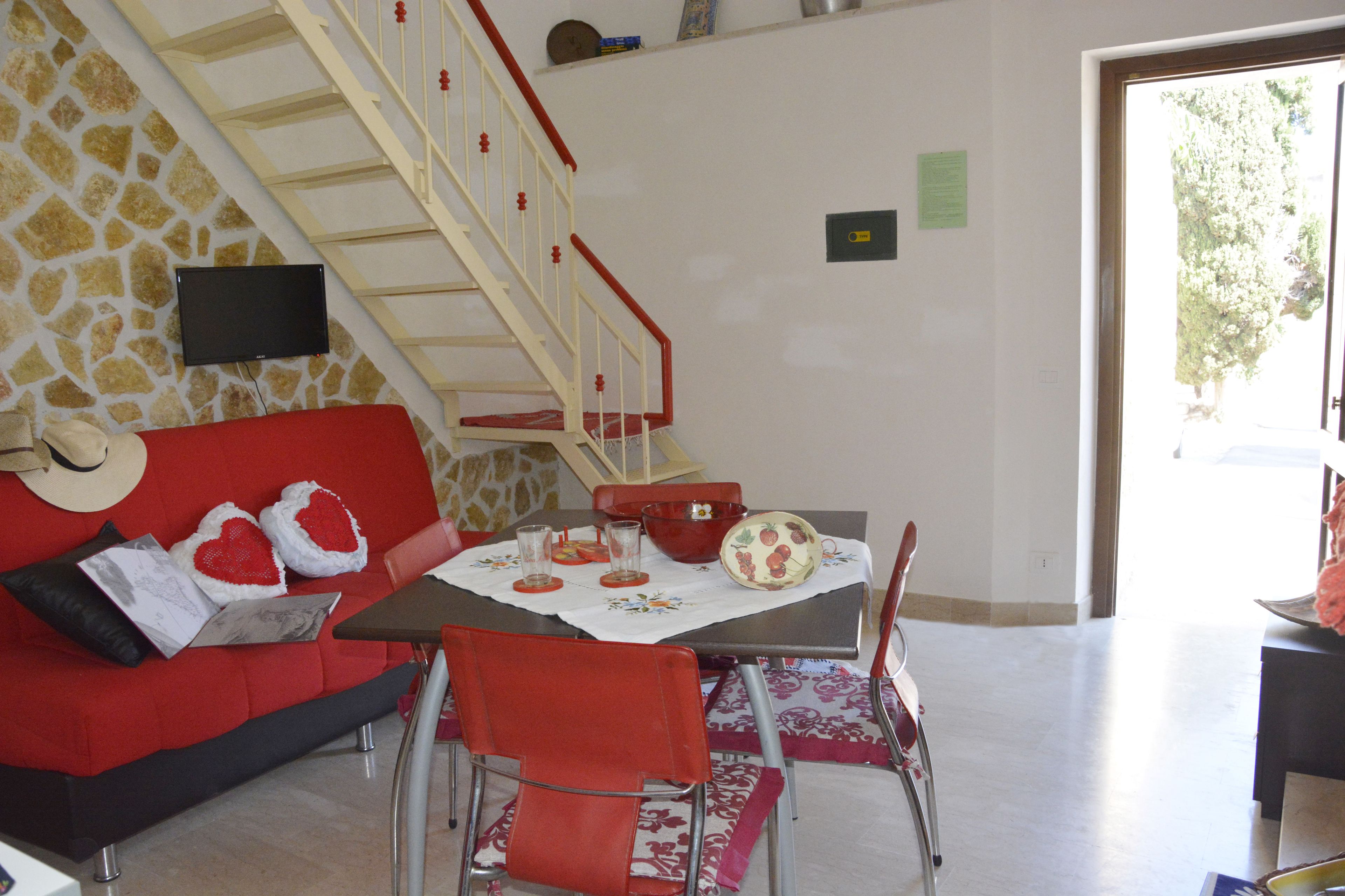 Un coin repas avec une table dressée pour deux personnes, un canapé avec des coussins rouges, un escalier et un mur en pierre.