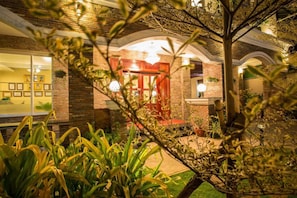 Um edifício de tijolos com uma porta vermelha, um candelabro e um jardim com plantas e uma árvore.