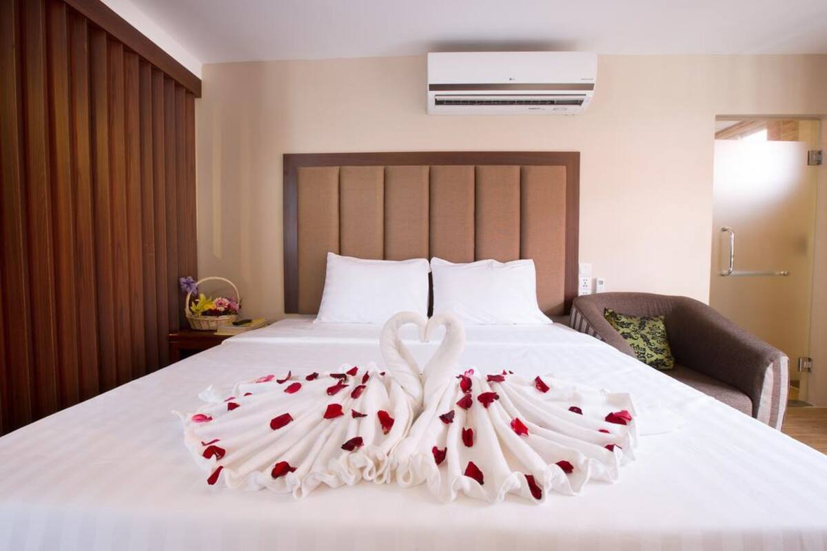 Een hotelkamer met een bed, een houten hoofdeinde, een bruine fauteuil en een tafeltje met een mand en bloemen.