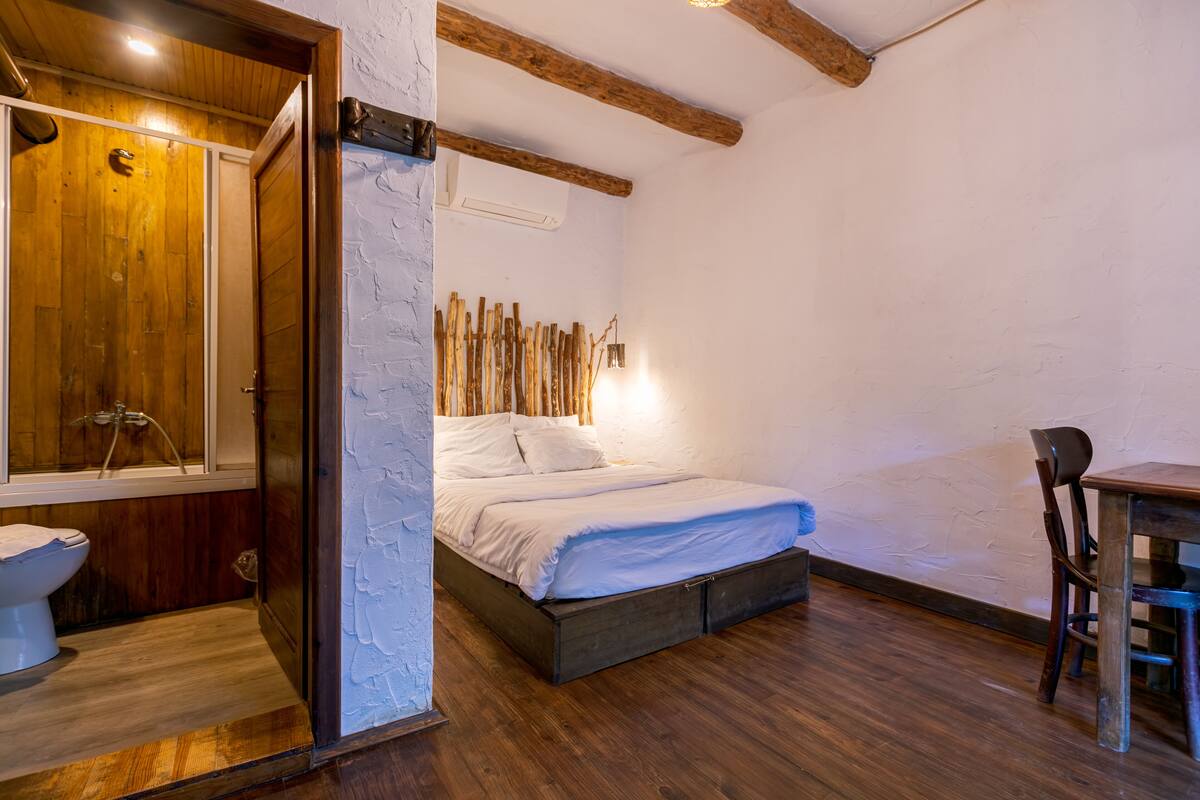 A bedroom with a wooden floor, a bed with white bedding, a wooden chair, and a wooden door leading to a bathroom.