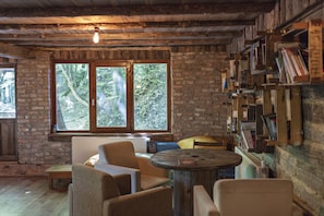 A rustic room with exposed brick walls, a large window with a view of trees, a round wooden table, and a bookshelf filled with books.