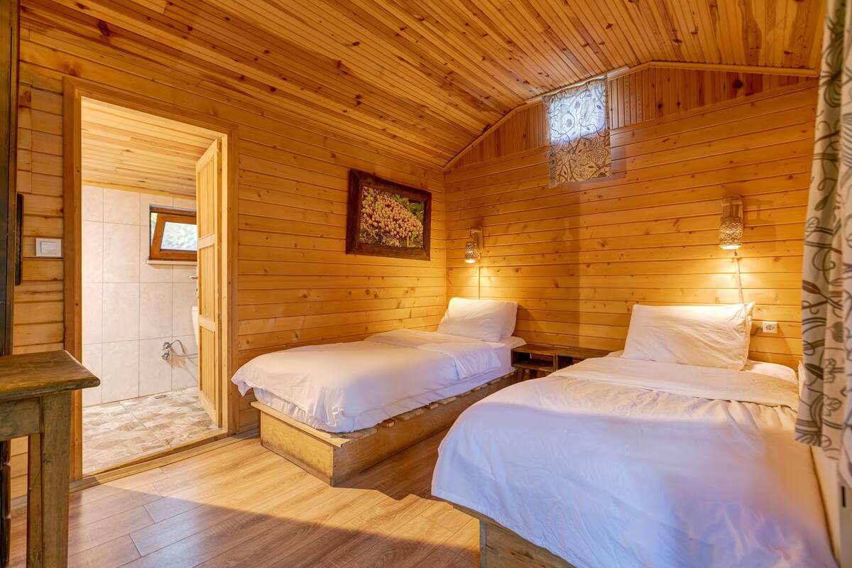 A wooden cabin room with two beds, a small table, a framed picture, and a window.