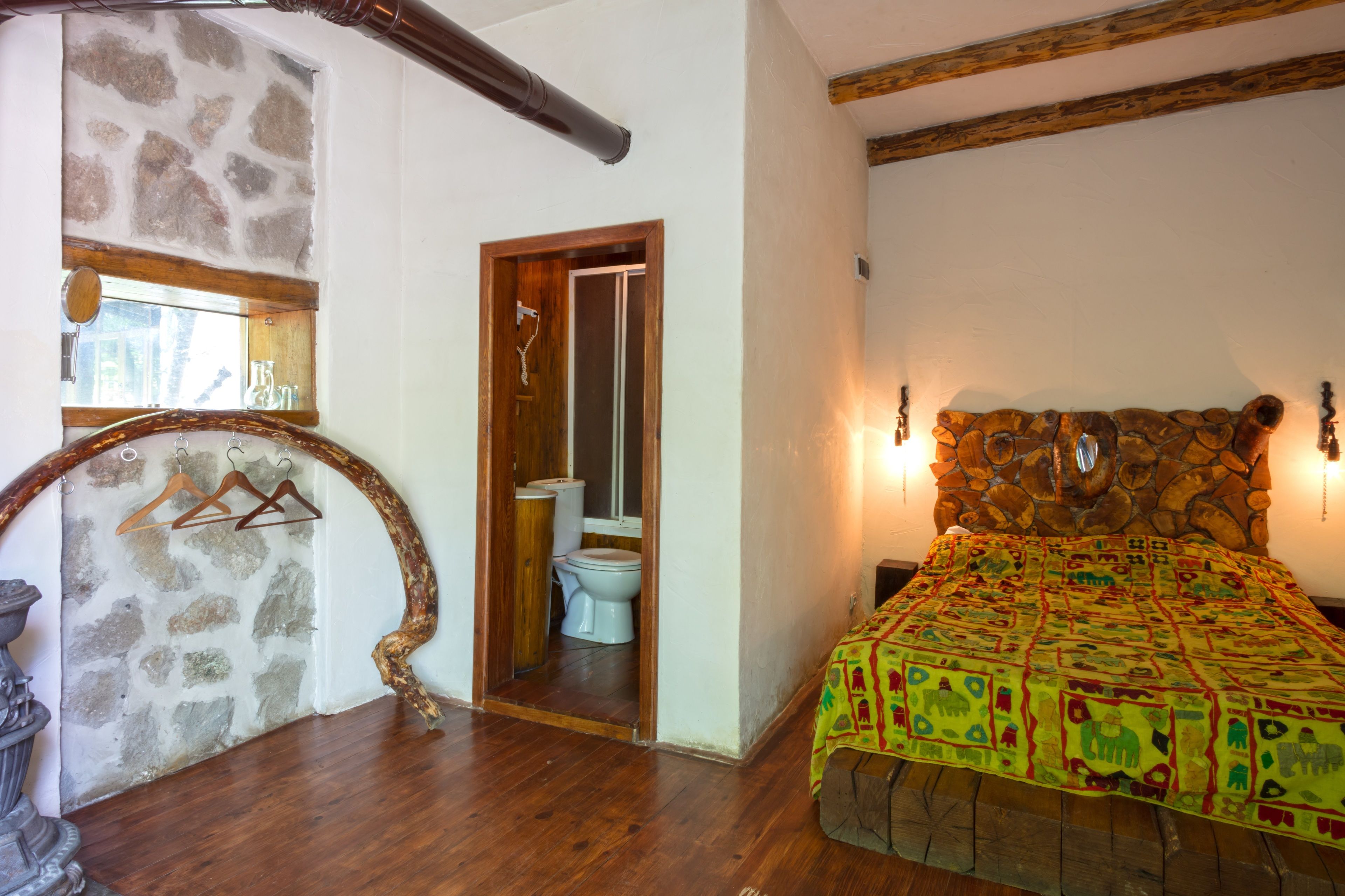 A bedroom with a stone wall, wooden floor, a bed with a colorful bedspread, a wooden door leading to a bathroom, and a stone archway.