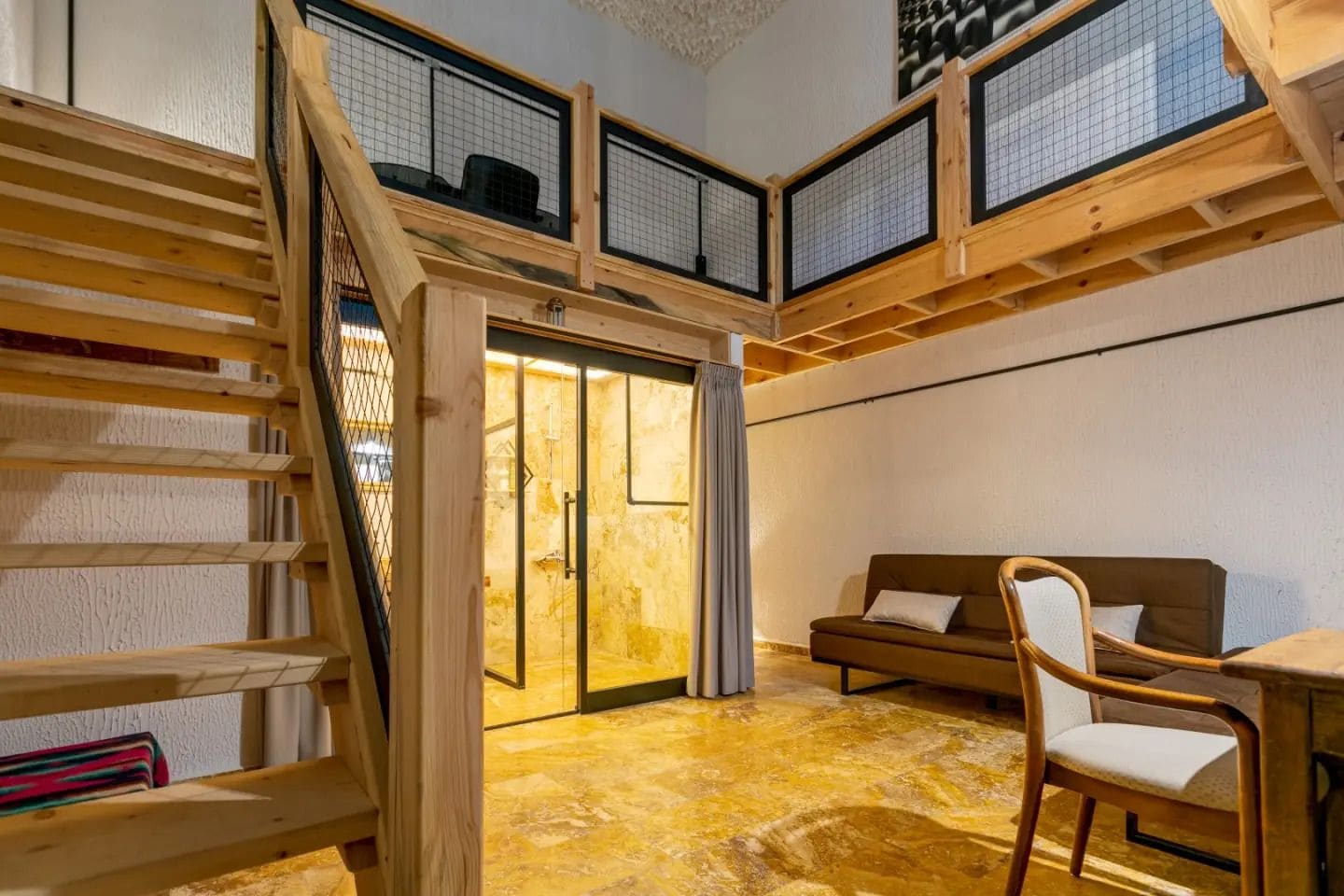 Una habitación con una escalera de madera, un sofá, una silla y un ventanal de vidrio que da a otro espacio.