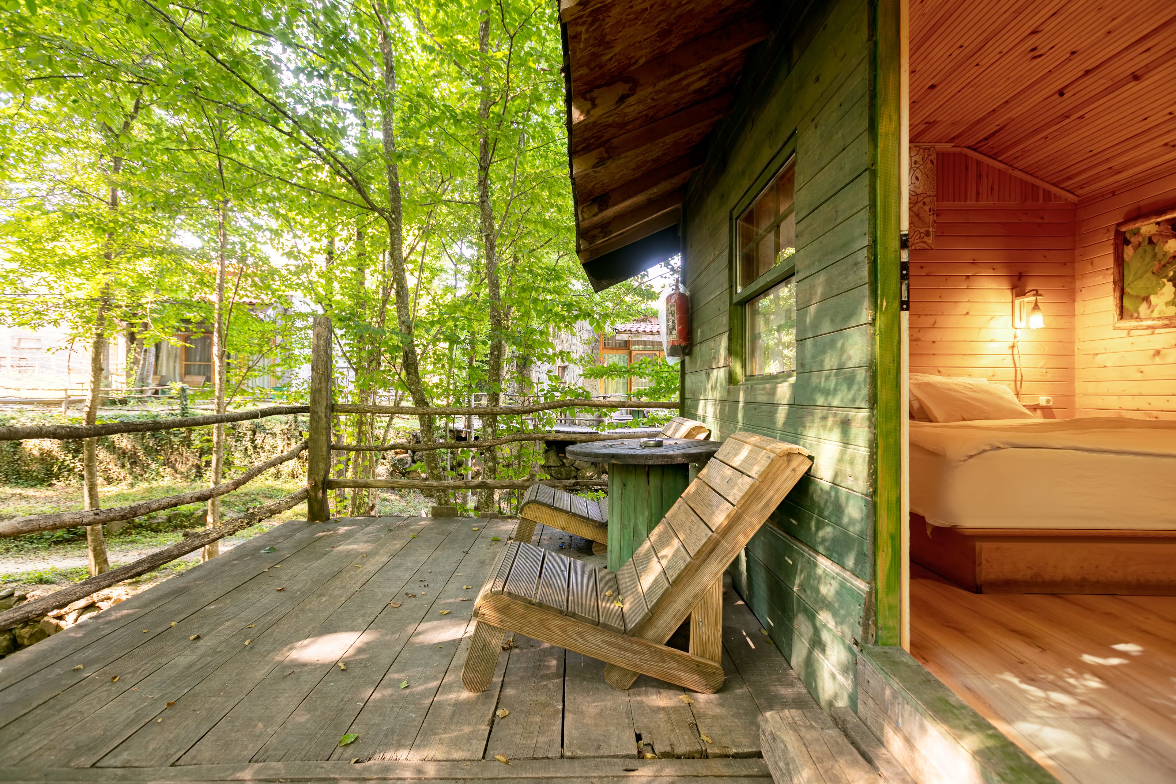 A wooden deck with a bench, a table, and a bed inside a cabin with a view of trees.